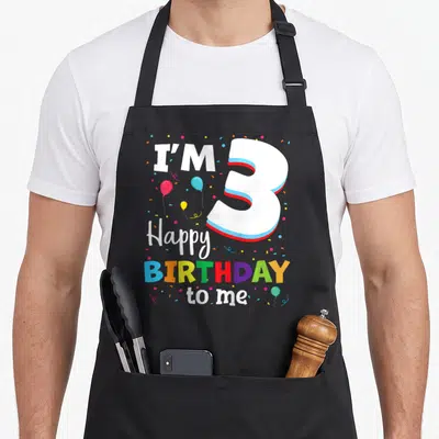 Three 3rd birthday apron - Cartoon number 3 with I'M Happy BIRTHDAY to me text, balloons and confetti in white red blue — shown on a black cotton canvas apron against a white background