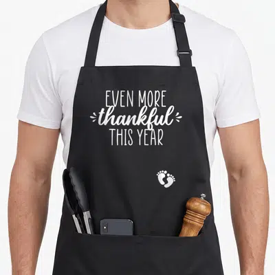 Announcement thankful thanksgiving apron - White text reading Even More Thankful This Year with baby footprints — shown on a black cotton canvas apron against a white background