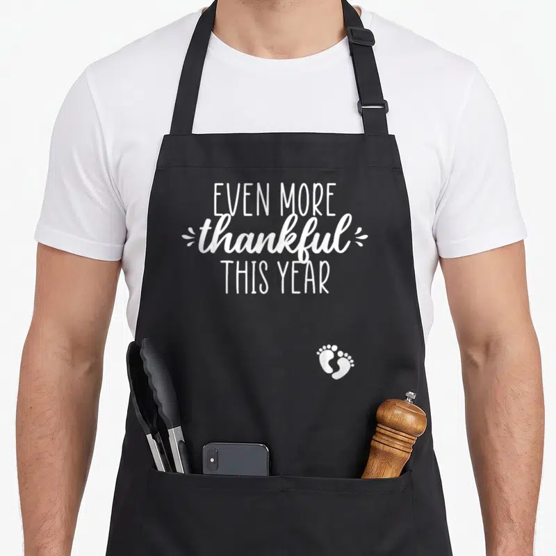 Announcement thankful thanksgiving apron - White text reading Even More Thankful This Year with baby footprints — shown on a black cotton canvas apron against a white background