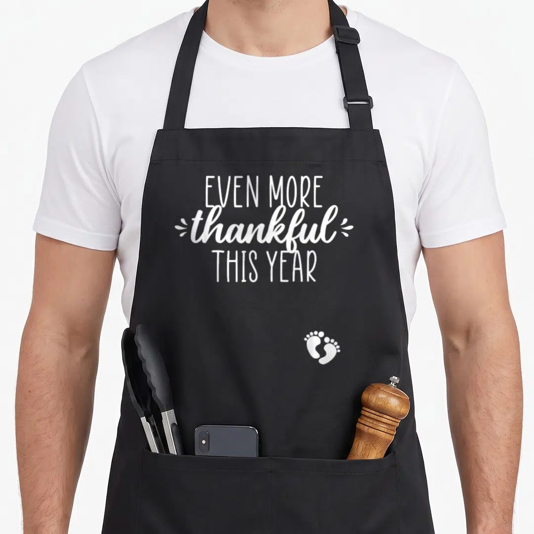 Announcement thankful thanksgiving apron - White text reading Even More Thankful This Year with baby footprints — shown on a black cotton canvas apron against a white background