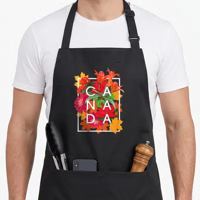 Flower canada pride apron - CANADA text over red orange flowers and yellow maple leaves in white frame — shown on a black cotton canvas apron against a white background