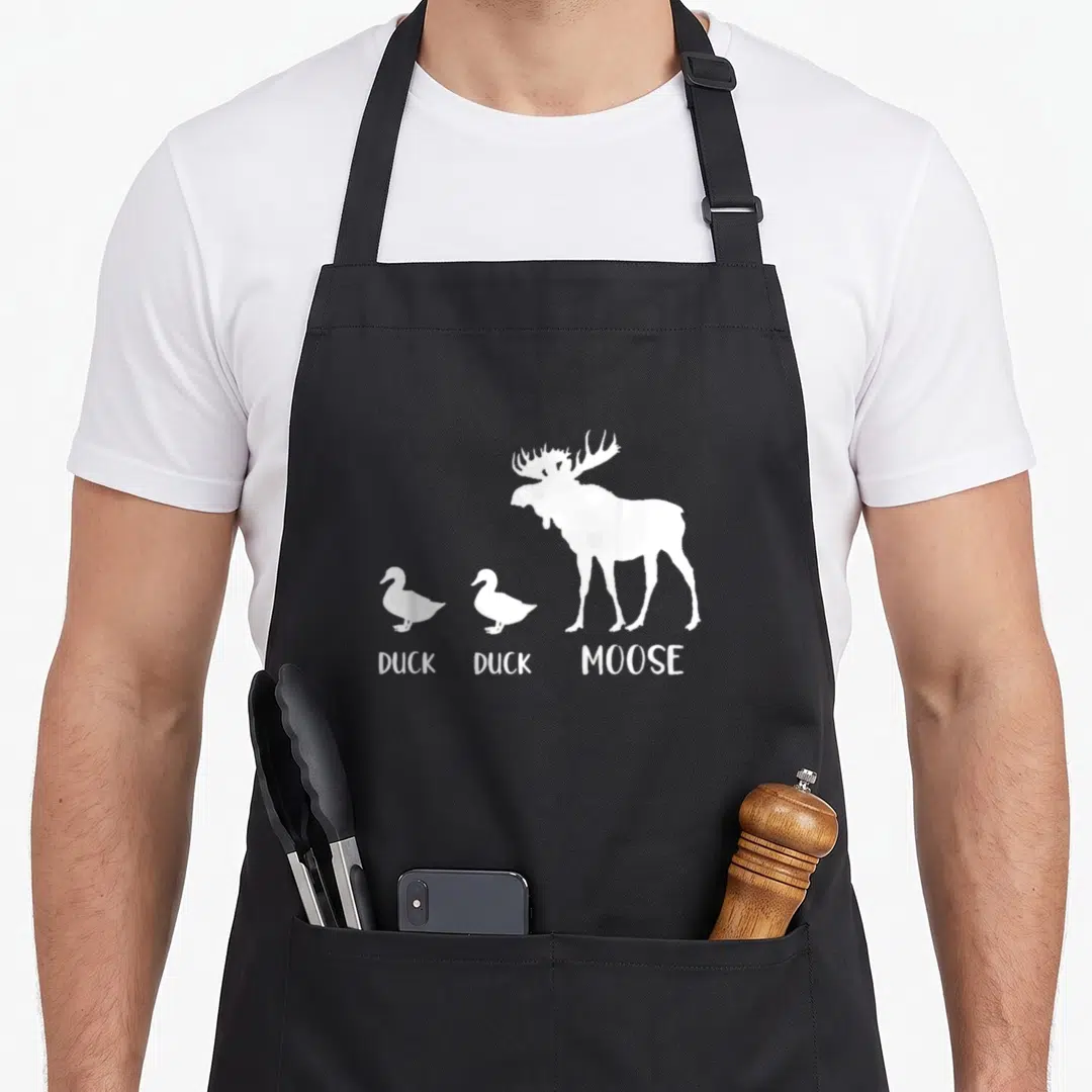 Moose duck apron - Three white animal silhouettes labeled DUCK DUCK MOOSE in a row — shown on a black cotton canvas apron against a white background