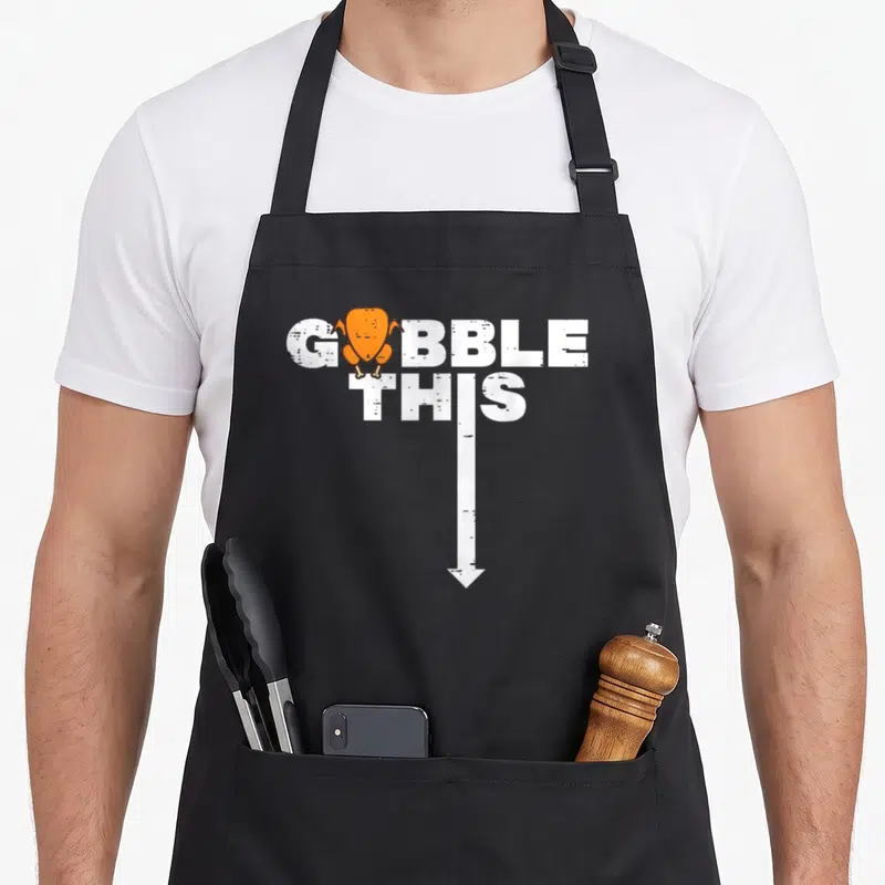 Gobble turkey thanksgiving apron - White text GOBBLE THIS with orange turkey and downward arrow — shown on a black cotton canvas apron against a white background