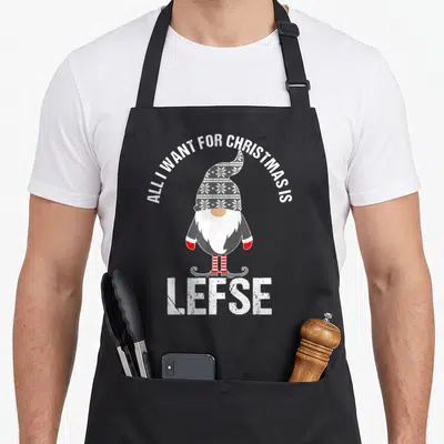 Tomte scandinavian aprons - Tomte gnome with grey snowflake hat and red striped socks under arched text and large LEFSE — shown on a black cotton canvas apron against a white background
