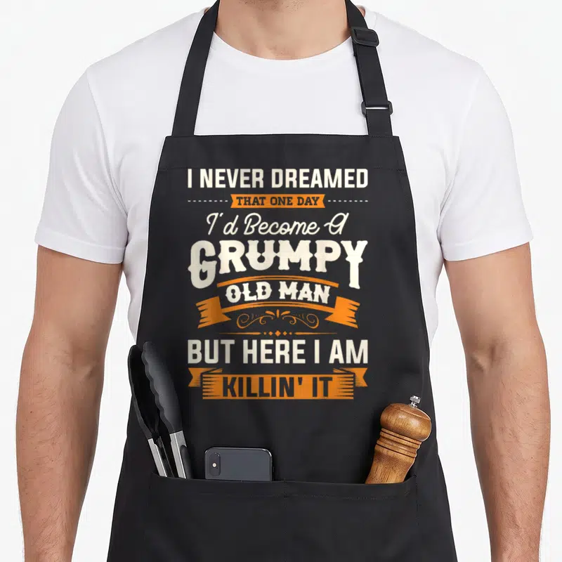 Dreamed grandpa apron - Stacked white text and orange ribbons reading I Never Dreamed That One Day Grumpy Old Man — shown on a black cotton canvas apron against a white background