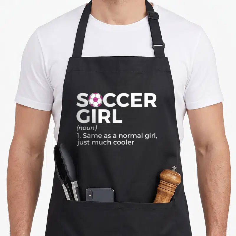 Definition soccer apron - SOCCER GIRL text with pink and white ball replacing the letter O in stacked typography — shown on a black cotton canvas apron against a white background