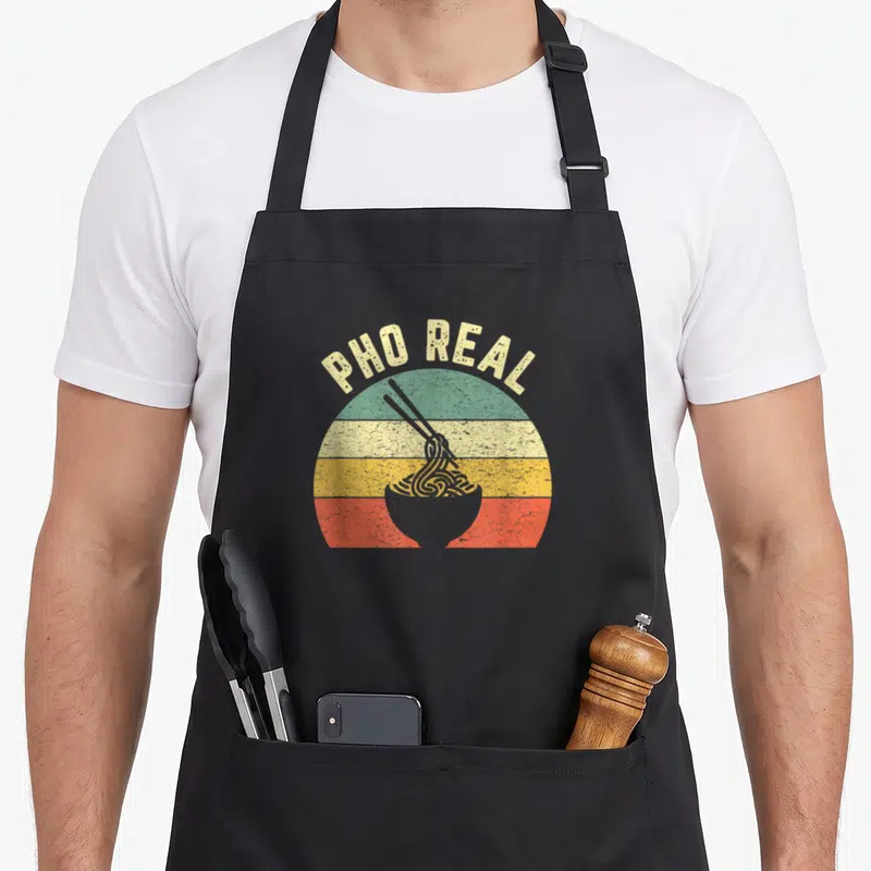 Noodle food aprons - PHO REAL arched text above striped circle and bowl of noodles with chopsticks — shown on a black cotton canvas apron against a white background