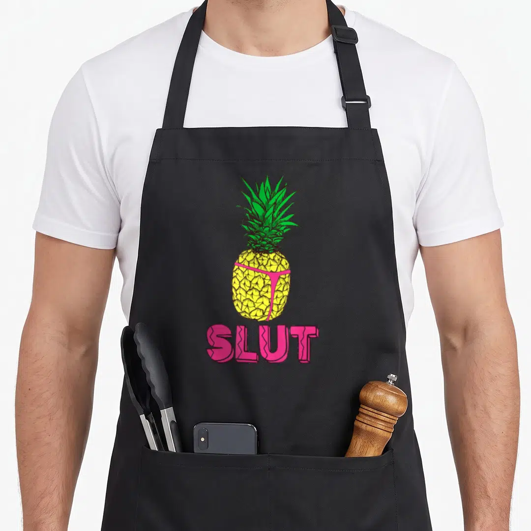 Pineapple tropical print aprons - Cartoon pineapple in pink lingerie above pink SLUT text on yellow and green — shown on a black cotton canvas apron against a white background
