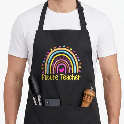 Future rainbow teacher apron - Rainbow with pink heart and Future Teacher text in yellow cartoon font — shown on a black cotton canvas apron against a white background