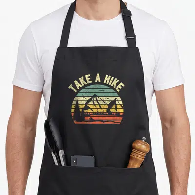 Hiker camping apron - TAKE A HIKE text arching over a striped sunset with mountain and bird silhouettes in retro colors — shown on a black cotton canvas apron against a white background