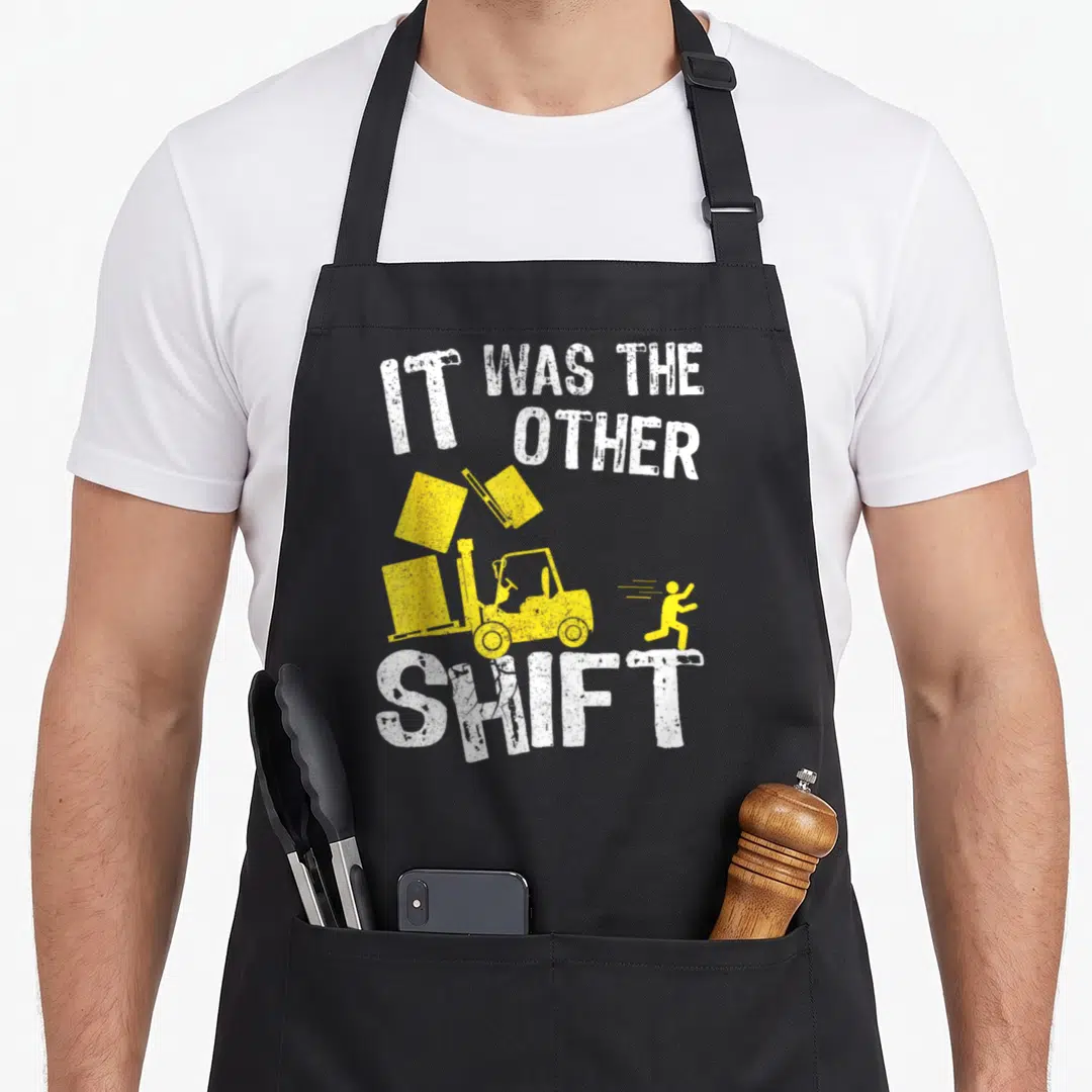 Shift forklift apron - Yellow forklift tipping three boxes with running figure and distressed white text — shown on a black cotton canvas apron against a white background