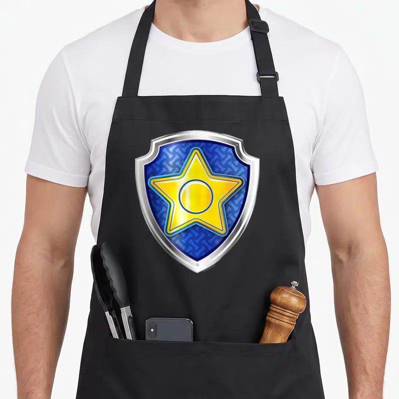 Funny badge chase birthday apron - Blue shield with yellow star and bone pattern enclosed by silver border — shown on a black cotton canvas apron against a white background