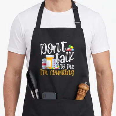 Talk pharmacy apron - Don't talk to me I'm counting text with orange and blue pill bottles — shown on a black cotton canvas apron against a white background