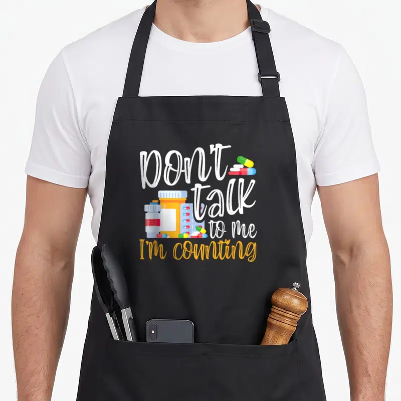 Talk pharmacy apron - Don't talk to me I'm counting text with orange and blue pill bottles — shown on a black cotton canvas apron against a white background