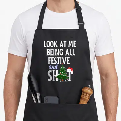 Festive xmas apron - Stacked text reading LOOK AT ME BEING ALL FESTIVE and SHIT with a green Christmas tree and red — shown on a black cotton canvas apron against a white background