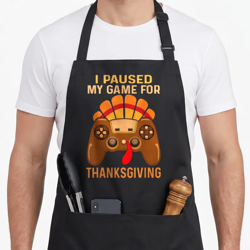 Gaming thanksgiving apron - I PAUSED MY GAME FOR THANKSGIVING text above a brown game controller turkey with orange — shown on a black cotton canvas apron against a white background