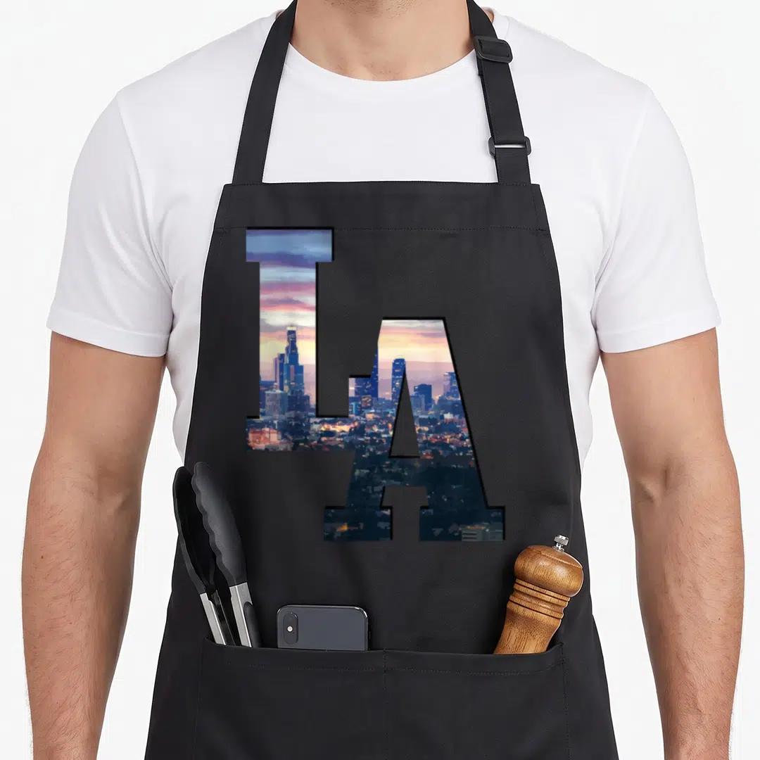 Skyline los angeles apron - LA letters filled with blue and orange city skyline at dusk — shown on a black cotton canvas apron against a white background