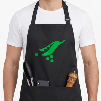 Cute peas apron - Green pea pod angled diagonally with scattered peas inside and outside the shell — shown on a black cotton canvas apron against a white background