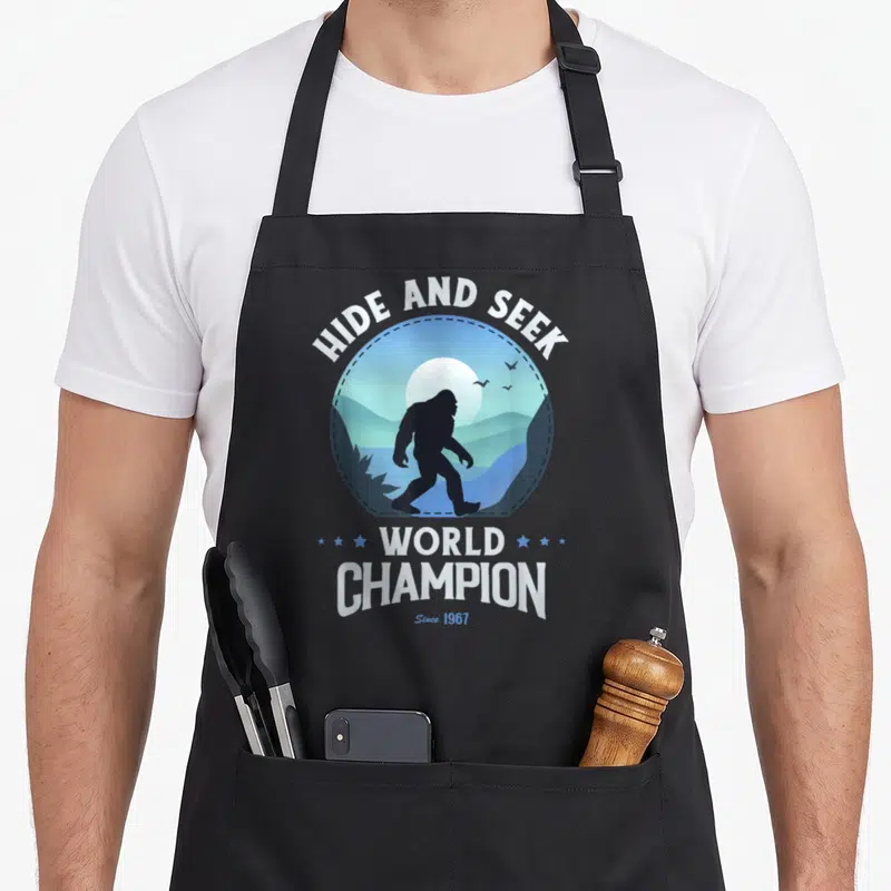 Retro seek bigfoot apron - Bigfoot silhouette in blue circle with HIDE AND SEEK WORLD CHAMPION text and stars — shown on a black cotton canvas apron against a white background