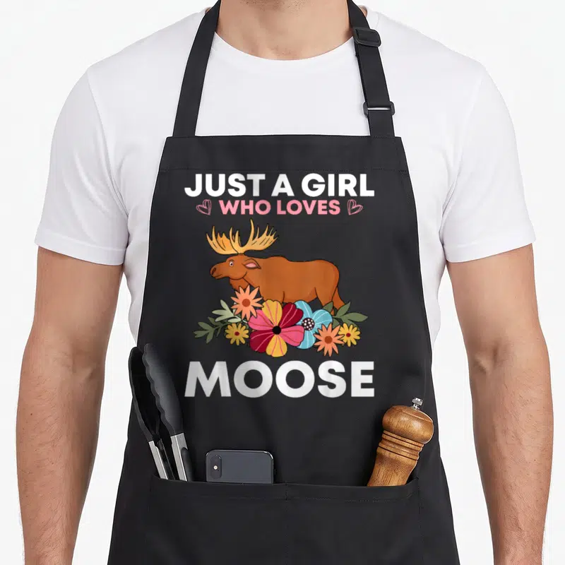 Antler animal apron - Cartoon moose with antlers surrounded by pink orange and blue flowers under white text — shown on a black cotton canvas apron against a white background