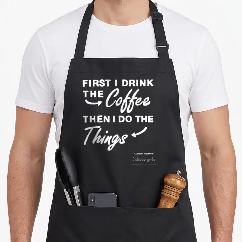 Show coffee apron - White stacked text reading FIRST I DRINK THE Coffee THEN I DO THE Things with arrows and signature — shown on a black cotton canvas apron against a white background