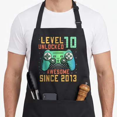 Level unlocked 10th birthday apron - Retro game controller in green and blue with orange text reading LEVEL 10 UNLOCKED — shown on a black cotton canvas apron against a white background