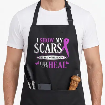 Ribbon domestic aprons - Purple and white text with awareness ribbon, butterfly, and heart symbol — shown on a black cotton canvas apron against a white background