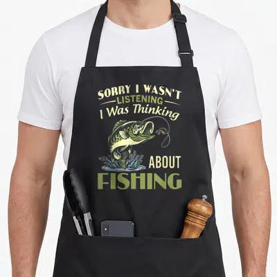 Wasnt fishing aprons - Cartoon jumping fish with blue splash and stacked beige green text saying Sorry I Wasn't — shown on a black cotton canvas apron against a white background