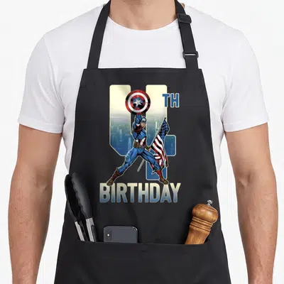 Cute 4th birthday apron - Captain America holding shield and flag before number 4 and skyline with TH BIRTHDAY text — shown on a black cotton canvas apron against a white background