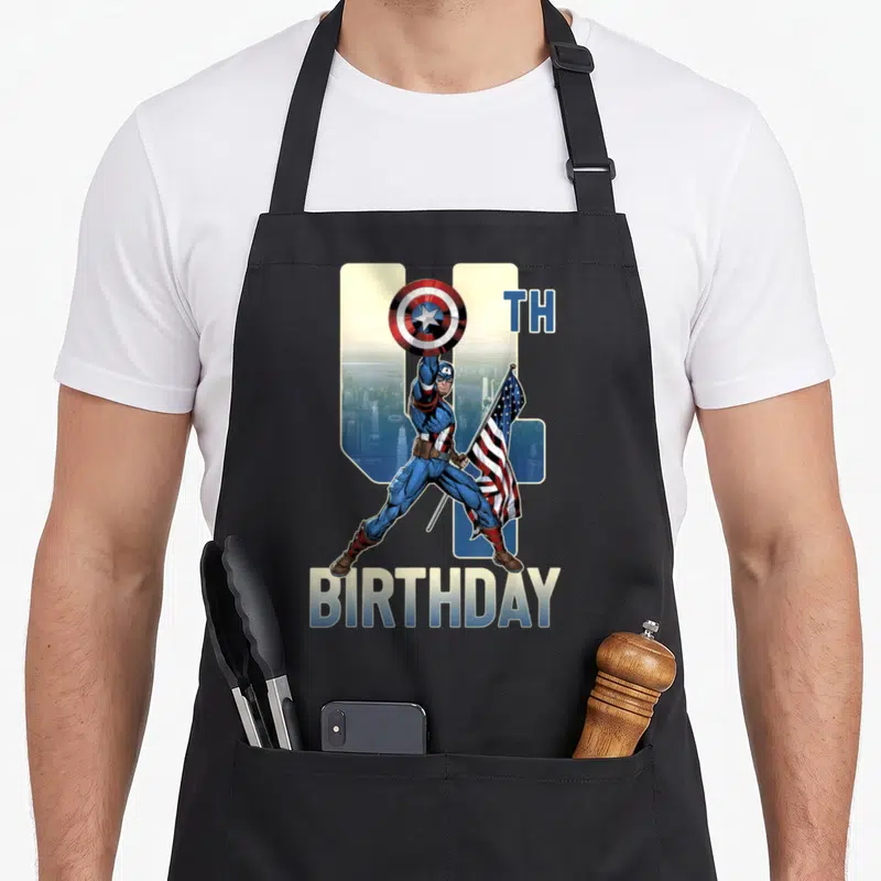Cute 4th birthday apron - Captain America holding shield and flag before number 4 and skyline with TH BIRTHDAY text — shown on a black cotton canvas apron against a white background