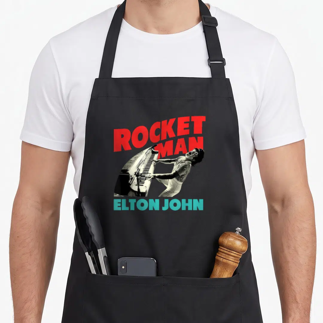 John piano apron - ROCKET MAN red text above man playing piano with ELTON JOHN teal text below — shown on a black cotton canvas apron against a white background