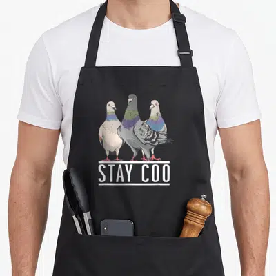Coo pigeon bird apron - Three cartoon pigeons in grey purple and green above white STAY COO text underlined by a bar — shown on a black cotton canvas apron against a white background