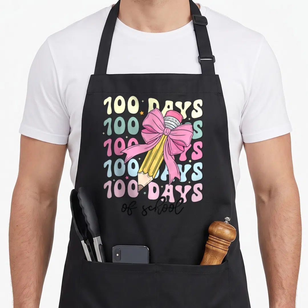 School bow apron - Yellow pencil with pink bow over five stacked 100 DAYS text lines in pastel colors — shown on a black cotton canvas apron against a white background