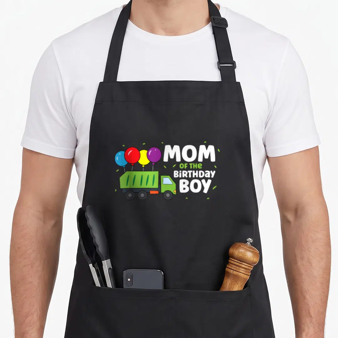 Birthday garbage truck apron - Green cartoon truck with four balloons and MOM OF THE BIRTHDAY BOY text — shown on a black cotton canvas apron against a white background