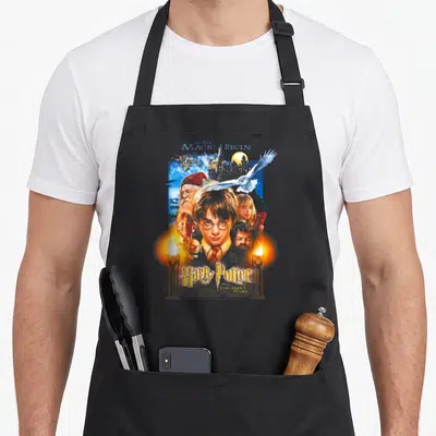 Movie novelty apron - Harry Potter collage with Let the Magic Begin text and two candles — shown on a black cotton canvas apron against a white background