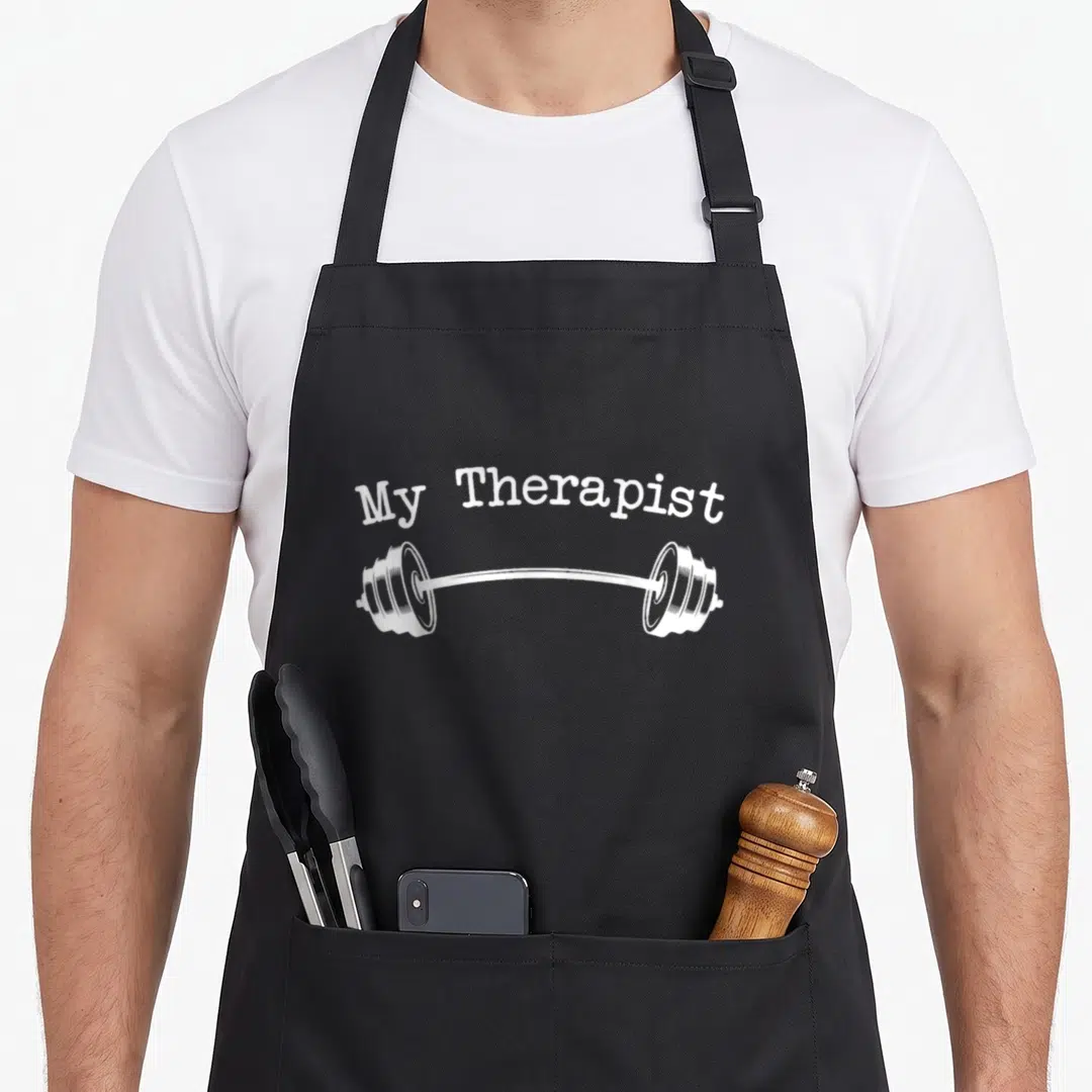 Therapist bodybuilding apron - White arched My Therapist text above a barbell illustration on black fabric — shown on a black cotton canvas apron against a white background