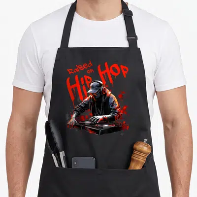 50th hip aprons - Raised on Hip Hop text above DJ with two turntables and red splatters — shown on a black cotton canvas apron against a white background