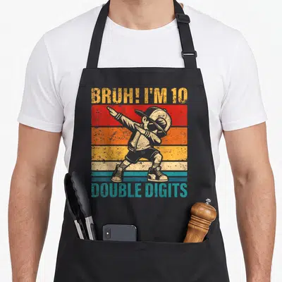 Bruh double digit birthday apron - Dabbing boy over five retro stripes with text BRUH I M 10 DOUBLE DIGITS — shown on a black cotton canvas apron against a white background