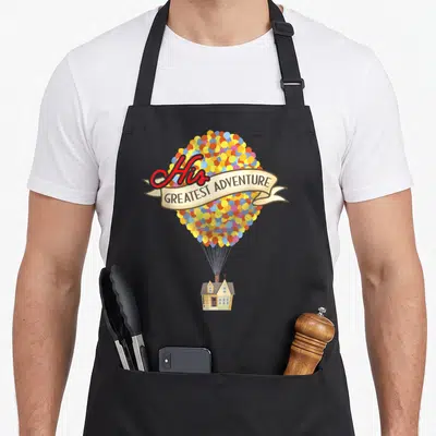 His novelty apron - House lifted by red yellow blue beige and brown balloons with His Greatest Adventure banner — shown on a black cotton canvas apron against a white background