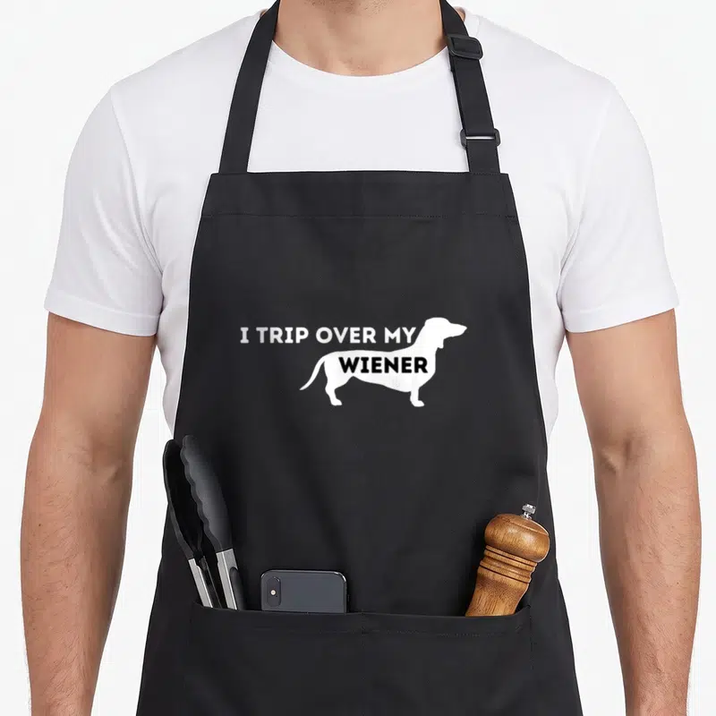 Trip badger apron - White dachshund silhouette with I TRIP OVER MY WIENER text inside body — shown on a black cotton canvas apron against a white background