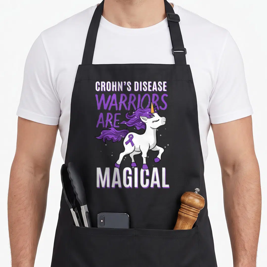 Crohn unicorn apron - White unicorn with purple mane and ribbon wearing warriors are magical text — shown on a black cotton canvas apron against a white background