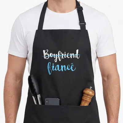 Engagement couple apron - White Boyfriend crossed out with blue line above blue fiancé script text — shown on a black cotton canvas apron against a white background