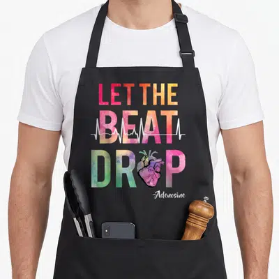 Beat nursing apron - Colorful Let The Beat Drop text with anatomical heart and white EKG line over grey background — shown on a black cotton canvas apron against a white background