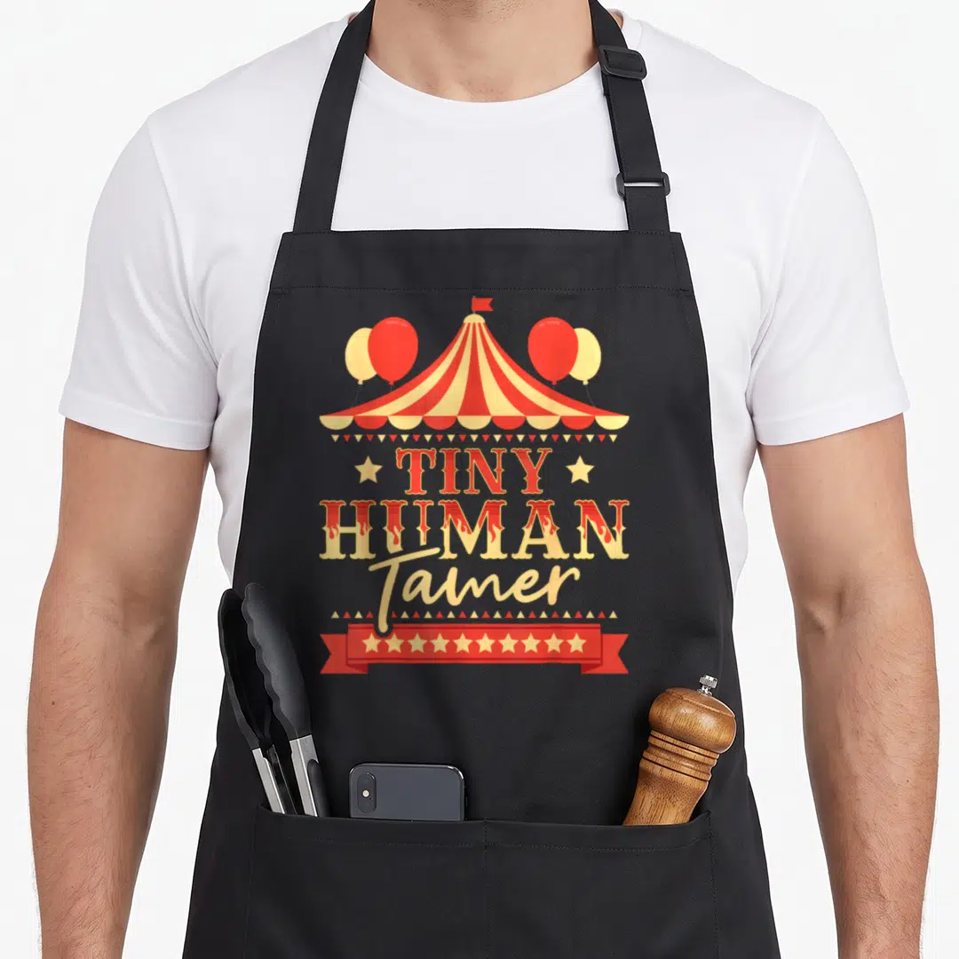 Tiny birthday apron - Circus tent with red balloons and TINY HUMAN Tamer text above a red ribbon banner — shown on a black cotton canvas apron against a white background