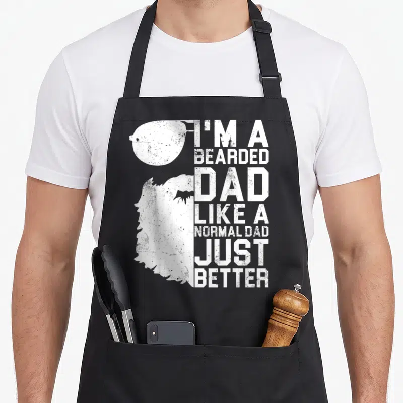 Bearded father day apron - White distressed text I'm a bearded dad like a normal dad just better next to sunglasses — shown on a black cotton canvas apron against a white background
