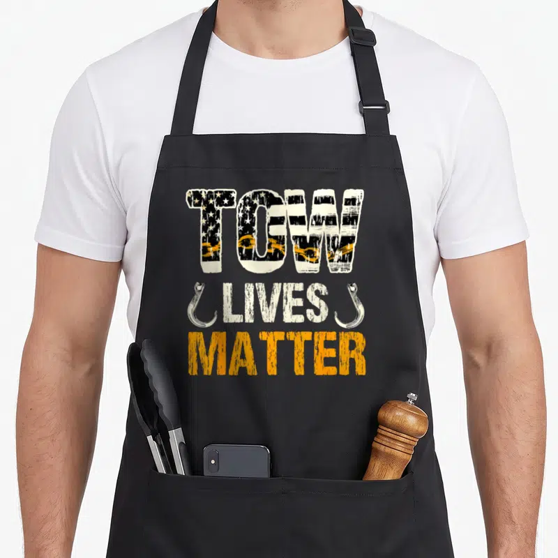 Hook truck apron - TOW LIVES MATRE text with flag pattern, two silver hooks, and orange chain links on black background — shown on a black cotton canvas apron against a white background