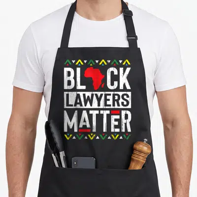 Month lawyer apron - Red Africa map as A in BLACK LAWYERS MATTER, distressed white text, colored triangle borders top — shown on a black cotton canvas apron against a white background
