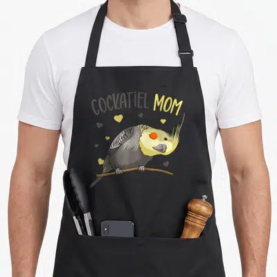Cute cockatiel bird apron - COCKATIEL MOM text above a perched cartoon bird with yellow and grey hearts on a branch — shown on a black cotton canvas apron against a white background