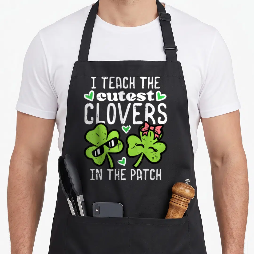 Teach st patrick day aprons - Two cartoon clovers with sunglasses and pink bow under white text I Teach The Cutest — shown on a black cotton canvas apron against a white background