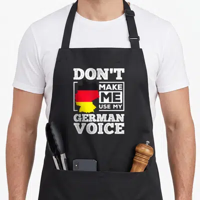 Use german apron - Distressed Germany map in black red yellow with text Don't Make Me Use My German Voice — shown on a black cotton canvas apron against a white background